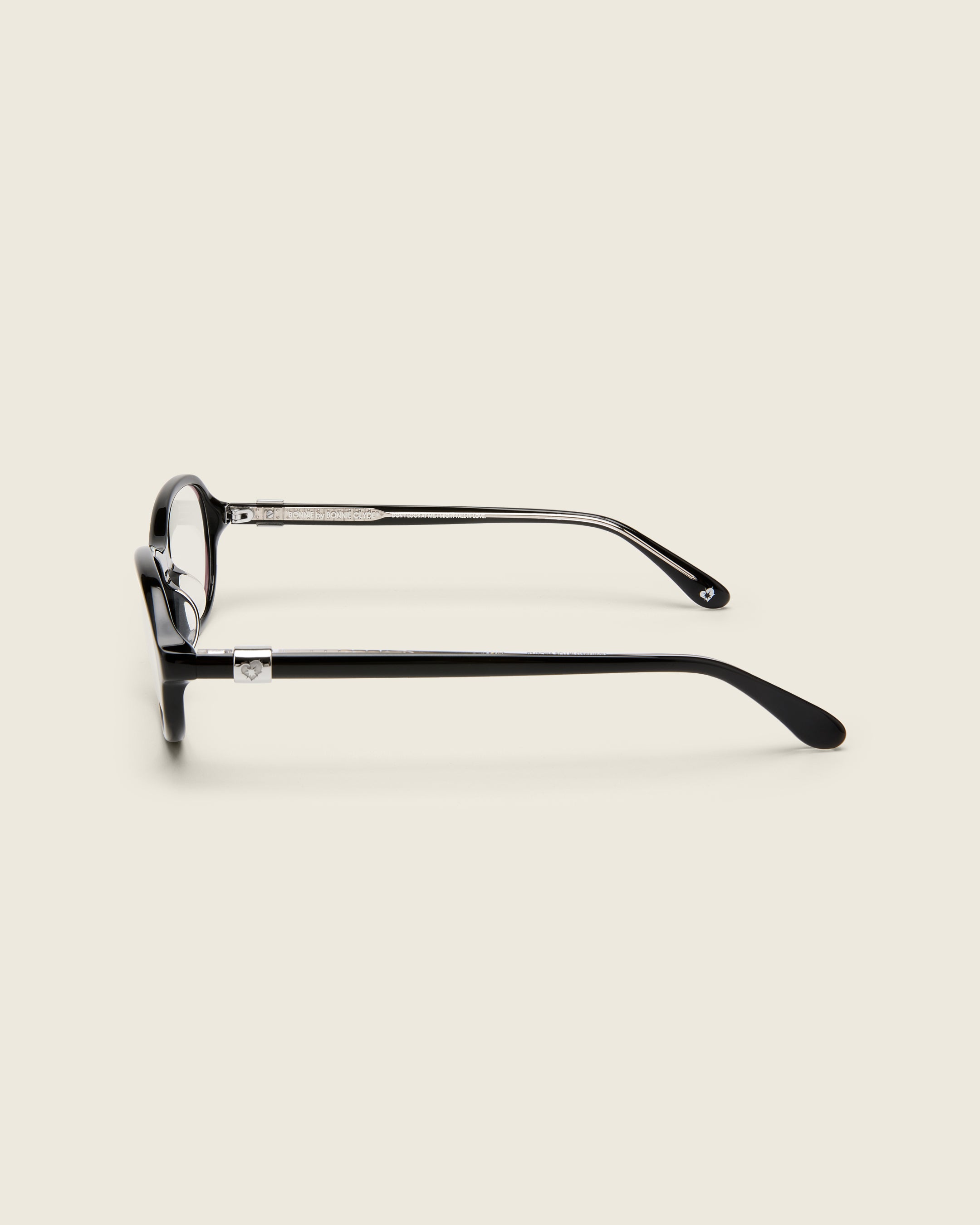 Rounded black acetate frames. Features raised browline curvature on a beige background. Features custom metal heart logo detail on arm temple. Side view.